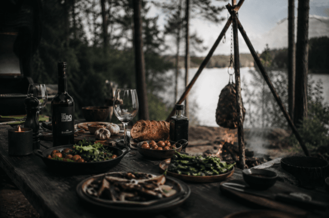 Rustic lakeside picnic with bread, salad, tomatoes, greens, wine, and a hanging roast over a campfire pit.
